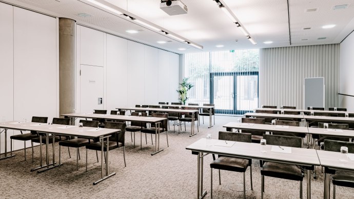 Modern meeting room with tables and chairs, a ceiling projector and large windows with blinds. Light, neutral colors dominate the room., &copy; Vienna House Easy Mo. Stuttgart