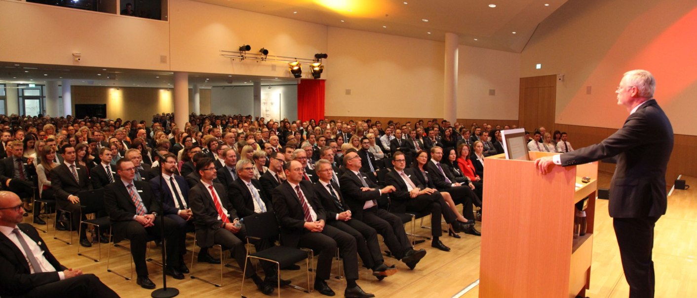 Ein Redner steht an einem Pult und spricht zu einem gro&szlig;en Publikum in einem modernen Konferenzsaal mit gelber Beleuchtung., &copy; ccbs