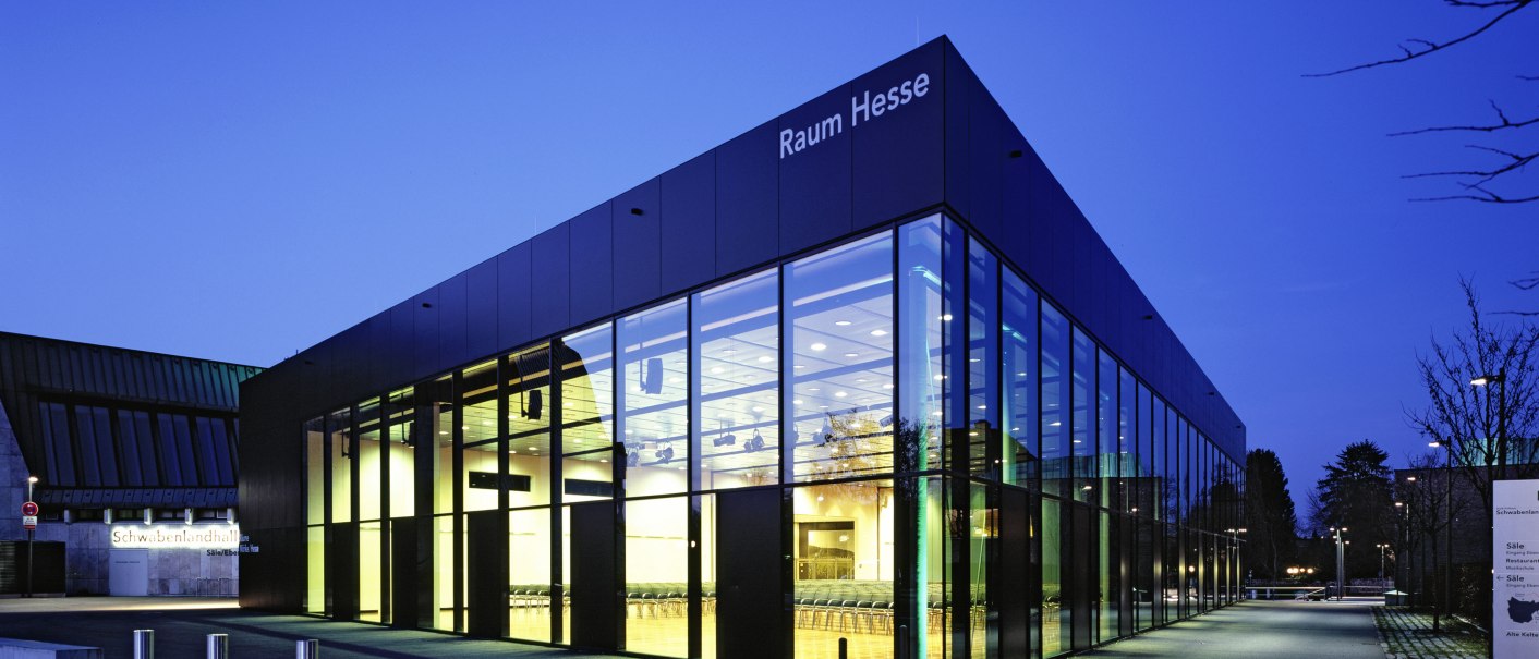 Die Schwabenlandhalle Fellbach bei Nacht. Moderne Glasfassade mit beleuchtetem Innenraum, umgeben von einem gepflasterten Weg und Bäumen., © Peter Hartung