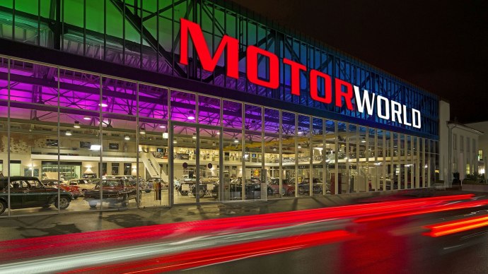 Die Motorworld Region Stuttgart bei Nacht, mit beleuchtetem Schriftzug und bunter Glasfront. Autos fahren vorbei und hinterlassen Lichtspuren., © MW Region Stuttgart Dieter Ege Die Motorworld Region Stuttgart bei Nacht, mit beleuchtetem Schriftzug und bunter Glasfront. Autos fahren vorbei und hinterlassen Lichtspuren., © MW Region Stuttgart Dieter Ege