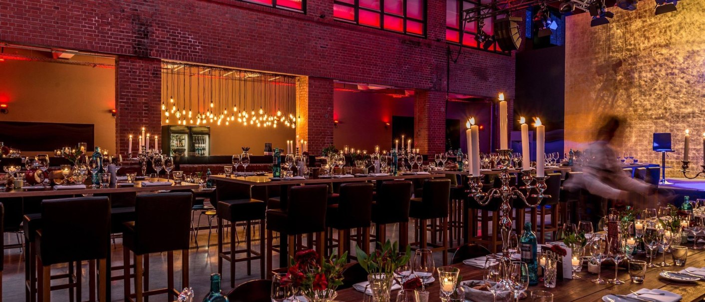 Eleganter Raum mit hohen Decken, gedeckten Tischen, Kerzenleuchtern und stimmungsvoller Beleuchtung in Rot und Gold., © rauschenbergercatering