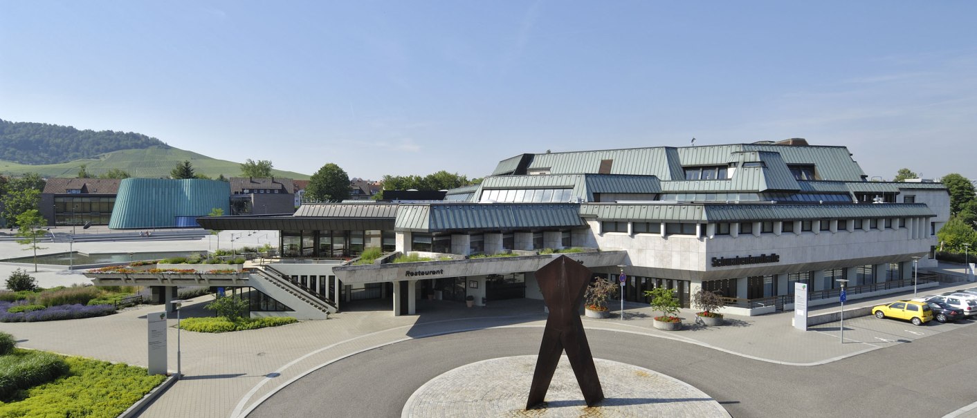 Die Schwabenlandhalle Fellbach mit moderner Architektur, umgeben von Grünflächen und einer Skulptur im Vordergrund. Im Hintergrund sind Hügel zu sehen., © Peter Hartung