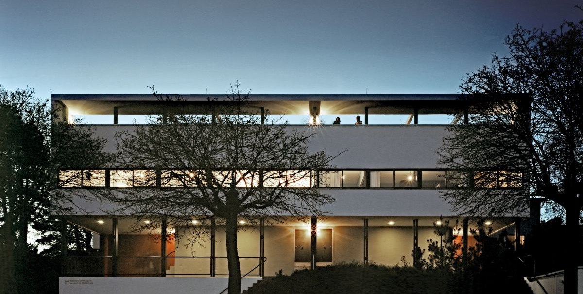 Modernes Gebäude der Weissenhofsiedlung bei Dämmerung, beleuchtet. Klare Linien und große Fenster prägen die Architektur., © Brigida Gonzalez