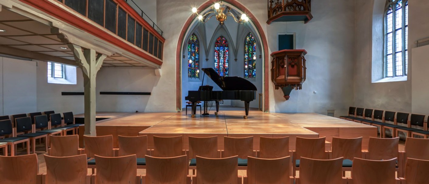 Interior view of the Kreuzkirche Nürtingen: Rows of chairs in front of a stage with a grand piano, colorful church windows in the background, wooden gallery and pulpit visible., © kkmaisch_gf Interior view of the Kreuzkirche Nürtingen: Rows of chairs in front of a stage with a grand piano, colorful church windows in the background, wooden gallery and pulpit visible., © kkmaisch_gf