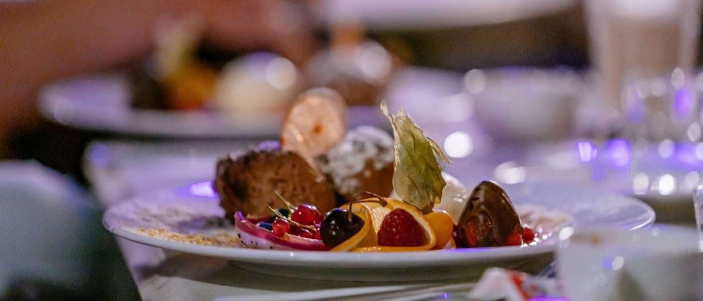 Ein kunstvoll angerichtetes Dessert mit Fr&uuml;chten, Schokolade und Dekoration auf einem Teller. Im Hintergrund unscharfe Lichter., &copy; Thomas Niederm&uuml;ller