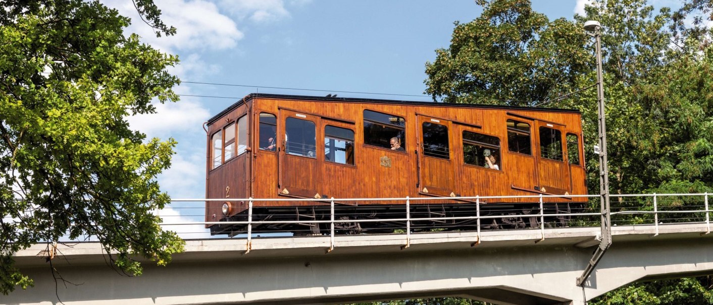 Eine historische Seilbahn mit Holzwagen fährt über eine Brücke, umgeben von grünen Bäumen und blauem Himmel., © TMBW, Gregor Lengler