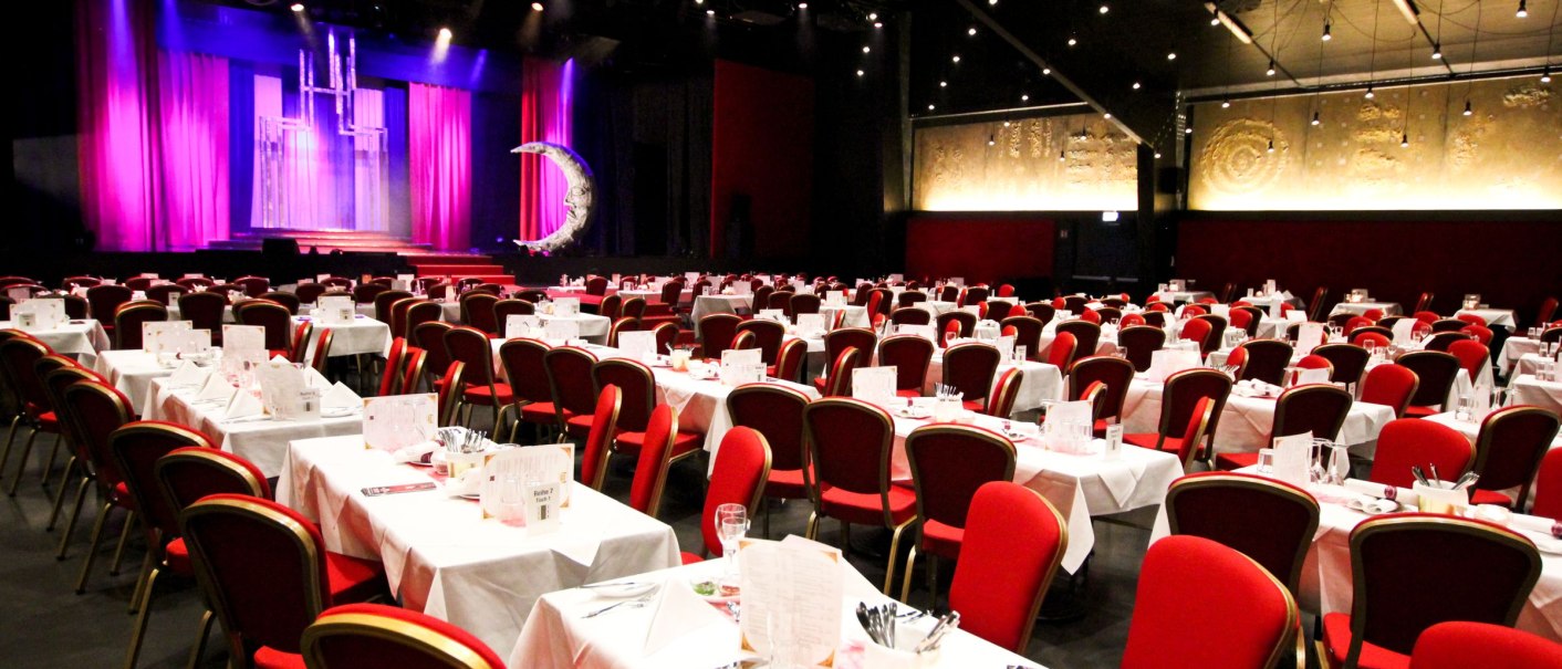 The Friedrichsbau Varieté Saal features a stage with purple curtains and set tables with red chairs in the foreground., © marta
