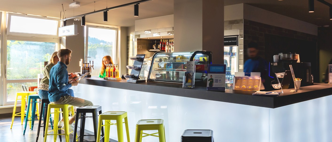 Moderne Bar mit beleuchtetem Tresen, bunten Hockern und Gästen, die Getränke genießen. Helle Fenster im Hintergrund., © a&o Hostels