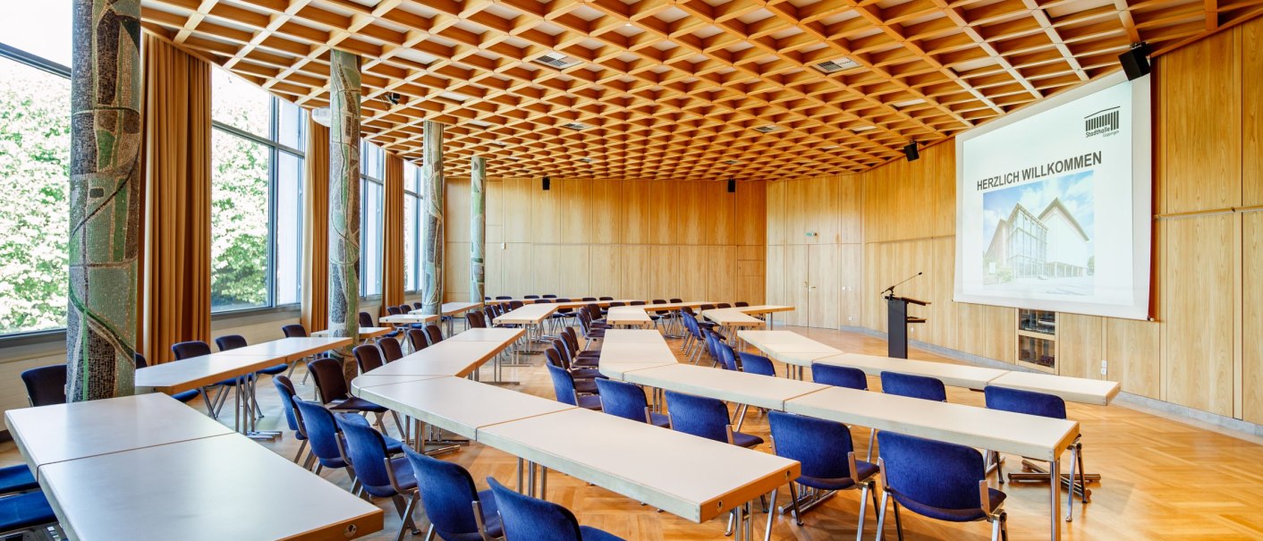 Der Märklinsaal in der Stadthalle Göppingen zeigt eine moderne Einrichtung mit Holzgitterdecke, blauen Stühlen und Tischen, sowie einer Leinwand mit Begrüßung., © Tobias Fröhner Photography