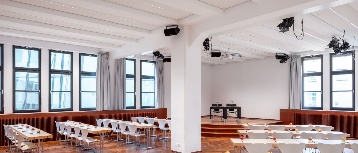 Großer Saal mit Tischen und Stühlen in parlamentarischer Anordnung, Holzboden, Bühne und großen Fenstern. An den Tischen liegen Äpfel und Gläser., © markus guhl fotografie