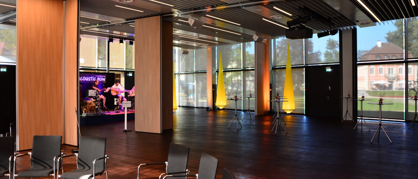 Moderner Saal mit Stühlen, Stehtischen und Bühne. Große Fenster lassen Tageslicht herein. Im Hintergrund spielt eine Band auf der Bühne., © Congress-Centrum Stadtgarten Schwäbisch Gmünd