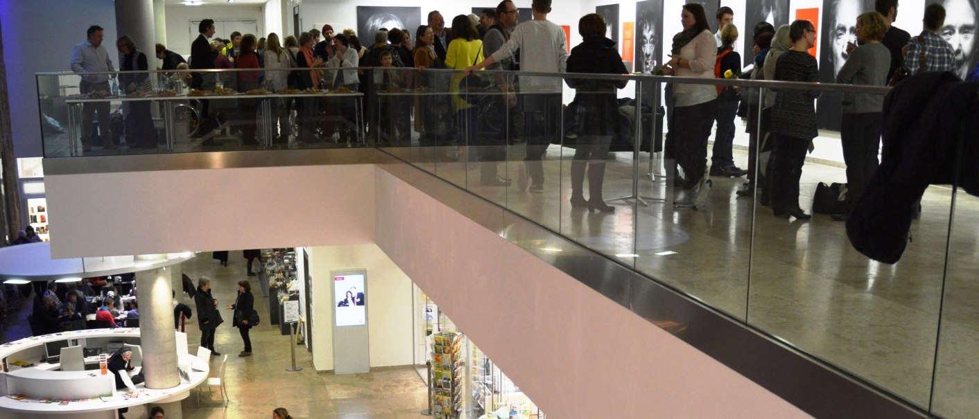 Eine Galerie mit Menschen auf zwei Ebenen; oben eine Ausstellung mit Porträts, unten ein Cafébereich., © hdkk_gf Eine Galerie mit Menschen auf zwei Ebenen; oben eine Ausstellung mit Porträts, unten ein Cafébereich., © hdkk_gf