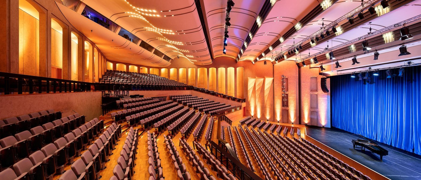Modern theater hall with blue curtains, empty rows of seats and warm light. The architecture is elegant and curved., © Stauch
