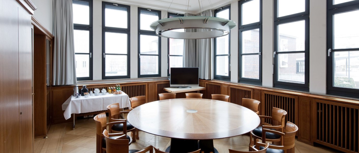 Ein heller Konferenzraum mit rundem Holztisch, Holzstühlen, großen Fenstern und einem Fernseher. Ein Tisch mit Getränken steht bereit., © Literaturhaus Stuttgart