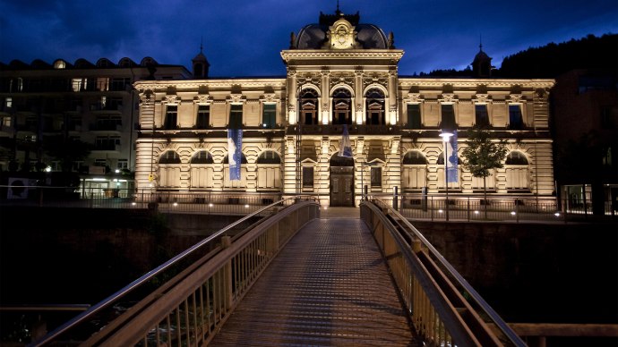 An illuminated building at night, the Forum K&ouml;nig-Karls-Bad, with a bridge in the foreground. The sky is dark blue., &copy; Forum Koenig-Karls-Bad