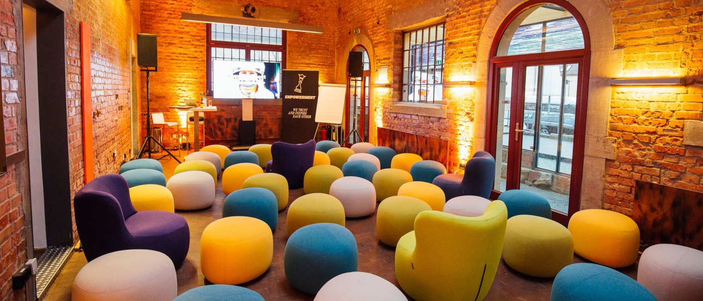 Ein Raum mit Backsteinwänden, bunten Sitzhockern und einem Bildschirm vorne. Die Beleuchtung ist warm und einladend., © Exometa Römerkastell