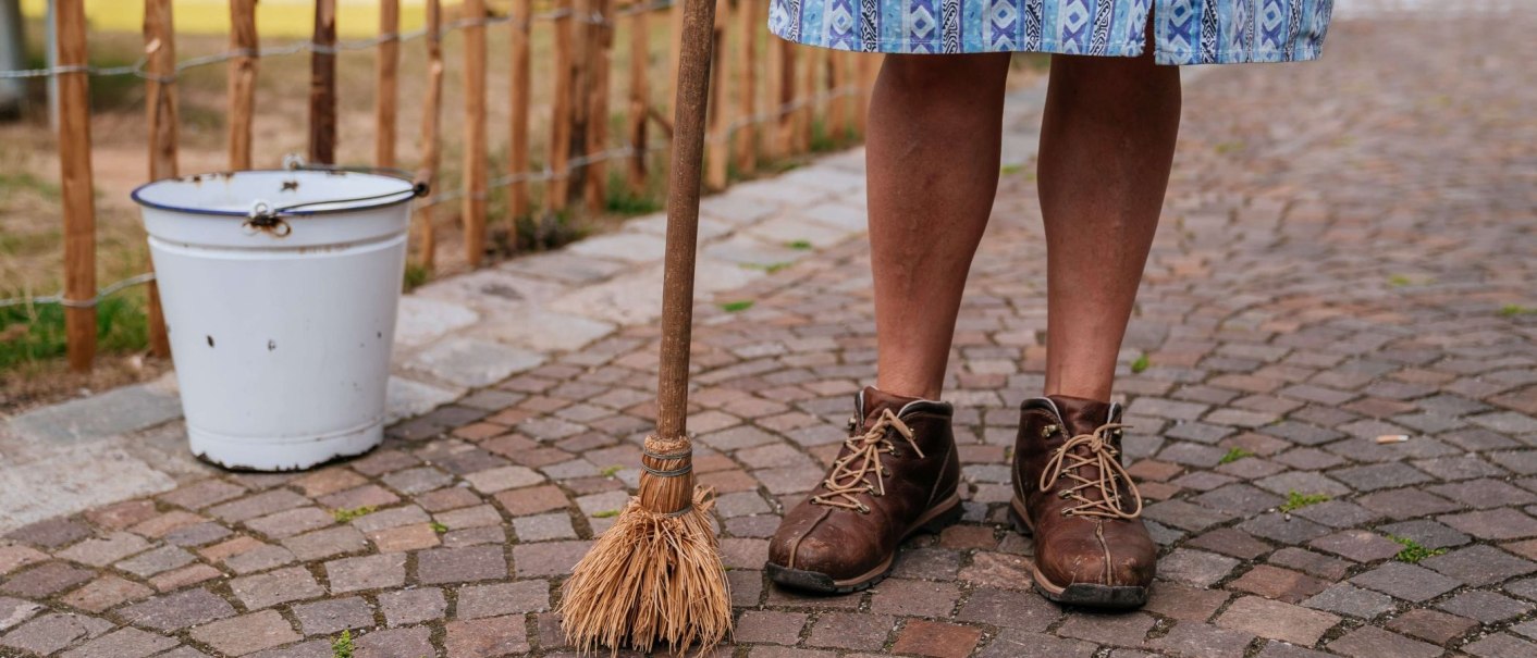 Person in braunen Schuhen steht auf Kopfsteinpflaster, hält einen Besen. Ein weißer Eimer steht daneben. Die Person trägt einen blauen Rock., © Thomas Niedermüller