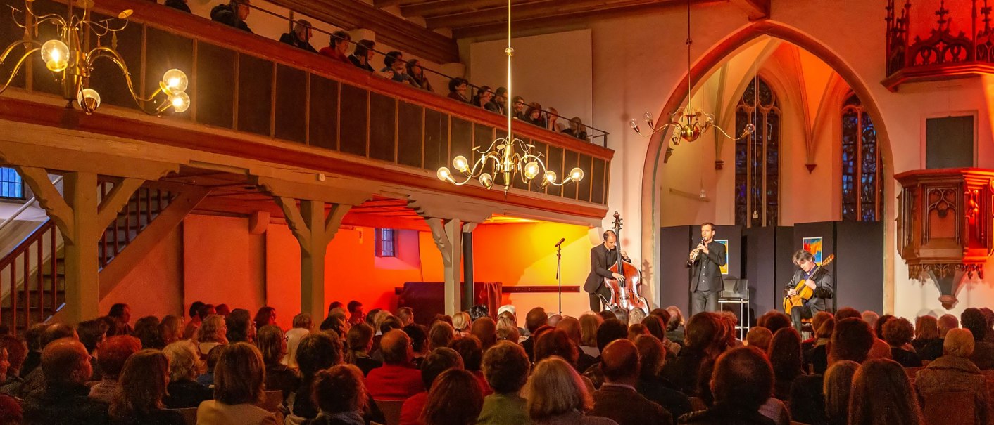 Concert in the Kreuzkirche Nürtingen with musicians on stage and a full audience. The church is warmly lit, with exposed wooden beams., © kkmaisch_gf Concert in the Kreuzkirche Nürtingen with musicians on stage and a full audience. The church is warmly lit, with exposed wooden beams., © kkmaisch_gf