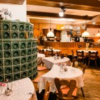 Cozy restaurant with wooden furniture, tablecloths and a green tiled stove. Lamps hanging from the ceiling, bar in the background., © Weinstube Kachelofen