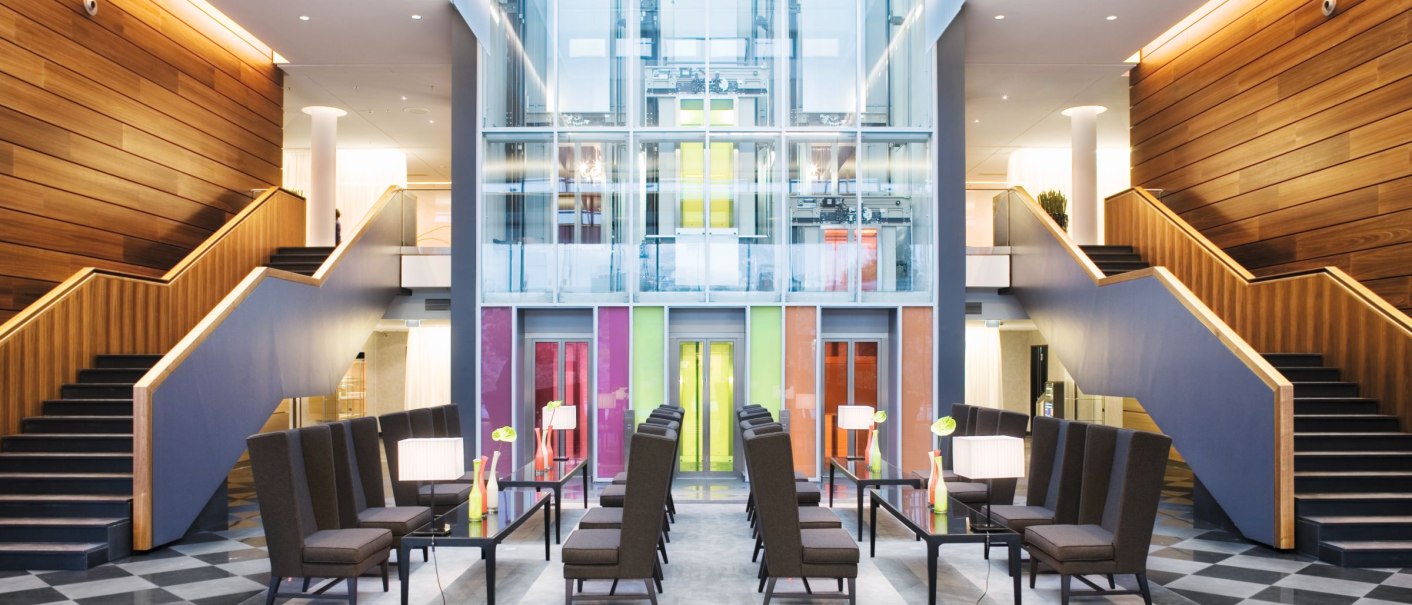 Moderne Lobby des Mövenpick Hotels Stuttgart Airport mit hohen Decken, symmetrischen Treppen und einem zentralen Sitzbereich., © Mövenpick Hotel Stuttgart Airport