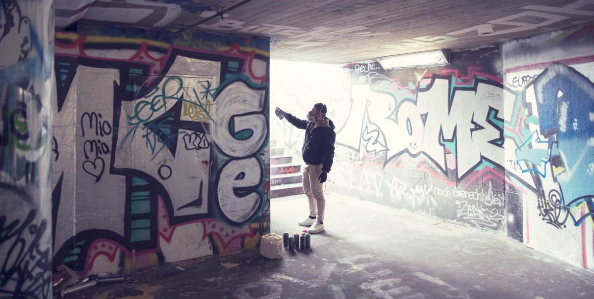 Eine Person sprüht Graffiti in einem Tunnel. Die Wände sind mit bunten Graffitis bedeckt. Auf dem Boden stehen Farbdosen., © hoponopo GmbH
