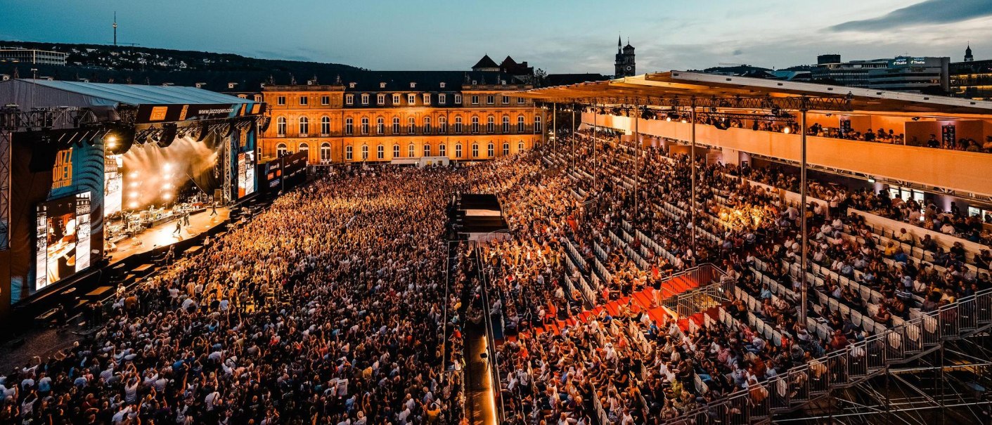 Large open-air concert at night with a crowd in front of an illuminated stage. Buildings and a tower can be seen in the background., © jazzopen stuttgart, Reiner Pfisterer Large open-air concert at night with a crowd in front of an illuminated stage. Buildings and a tower can be seen in the background., © jazzopen stuttgart, Reiner Pfisterer