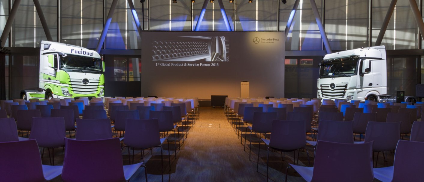 Empty conference room with rows of chairs, two trucks and a screen with the Mercedes-Benz logo. Lighting and modern architecture characterize the room., &copy; Carl Benz Arena