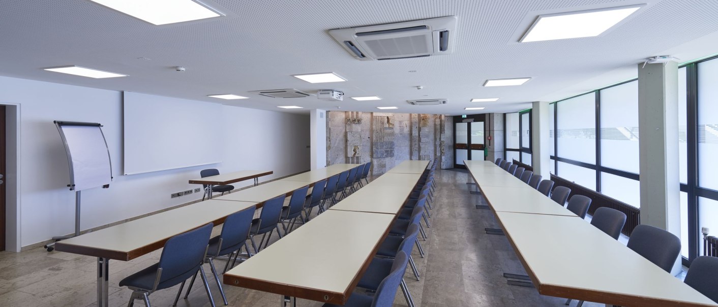 Modern conference room with long tables, blue chairs, flipchart and projector. Large windows on the right-hand side., © Peter Hartung Modern conference room with long tables, blue chairs, flipchart and projector. Large windows on the right-hand side., © Peter Hartung