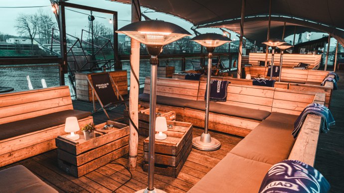 Comfortable seating on a covered upper deck with wooden benches, radiant heaters and blankets. A river can be seen in the background., © Fridas Pier