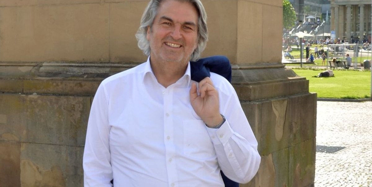 Ein Mann in weißem Hemd steht lächelnd vor einem historischen Gebäude auf einem sonnigen Platz., © Wolfgang Seljé Ein Mann in weißem Hemd steht lächelnd vor einem historischen Gebäude auf einem sonnigen Platz., © Wolfgang Seljé