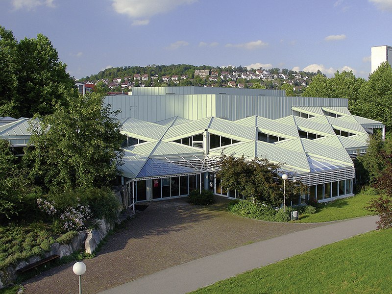 Die Stadthalle Leonberg mit ihrem markanten, geometrischen Dachdesign, umgeben von Bäumen und Grünflächen, bei sonnigem Wetter., © Stadthalle Leoonberg