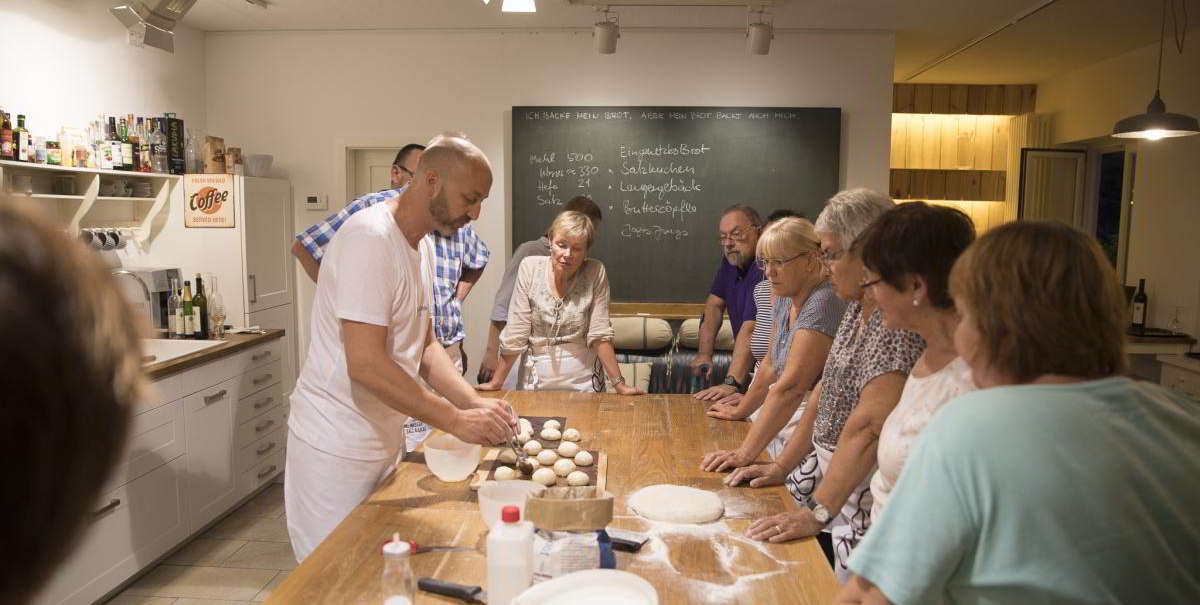 Eine Gruppe von Menschen steht um einen Tisch in einer Küche und lernt, wie man Brezeln backt. Ein Mann zeigt den Teilnehmern den Teig., © hoponopo GmbH Eine Gruppe von Menschen steht um einen Tisch in einer Küche und lernt, wie man Brezeln backt. Ein Mann zeigt den Teilnehmern den Teig., © hoponopo GmbH