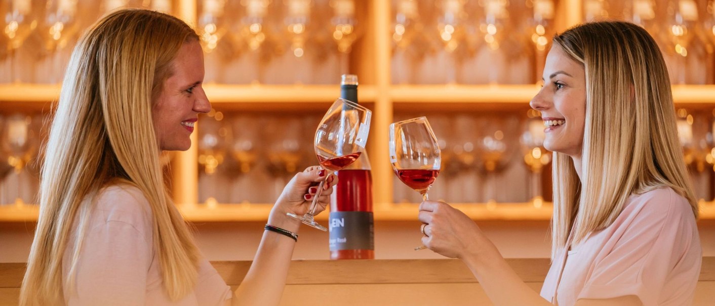 Zwei Frauen stoßen mit Weingläsern an, lächelnd in einem Weinbaumuseum. Im Hintergrund sind Weinflaschen und Gläser zu sehen., © Thomas Niedermüller Zwei Frauen stoßen mit Weingläsern an, lächelnd in einem Weinbaumuseum. Im Hintergrund sind Weinflaschen und Gläser zu sehen., © Thomas Niedermüller