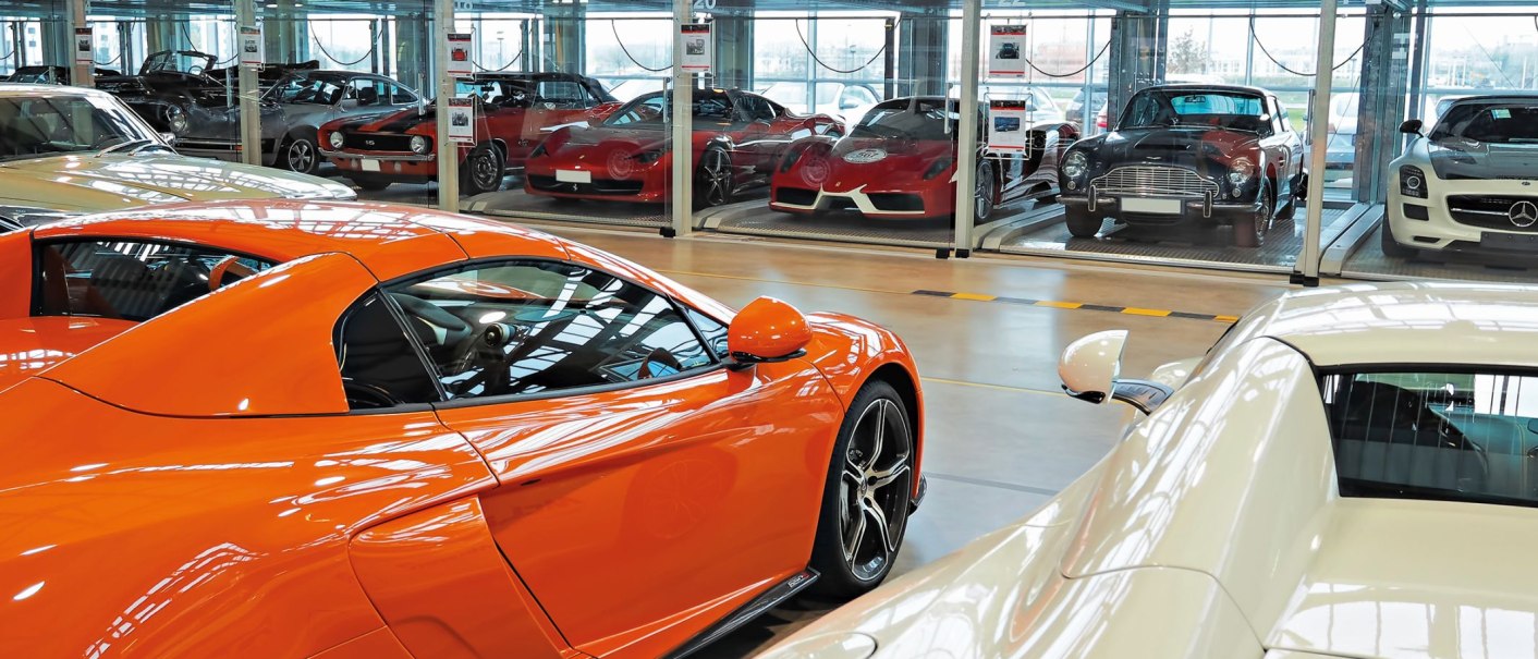 Several luxury cars in glass boxes at Motorworld Region Stuttgart. An orange car in the foreground, other vehicles in the background., © MW Region Stuttgart Several luxury cars in glass boxes at Motorworld Region Stuttgart. An orange car in the foreground, other vehicles in the background., © MW Region Stuttgart