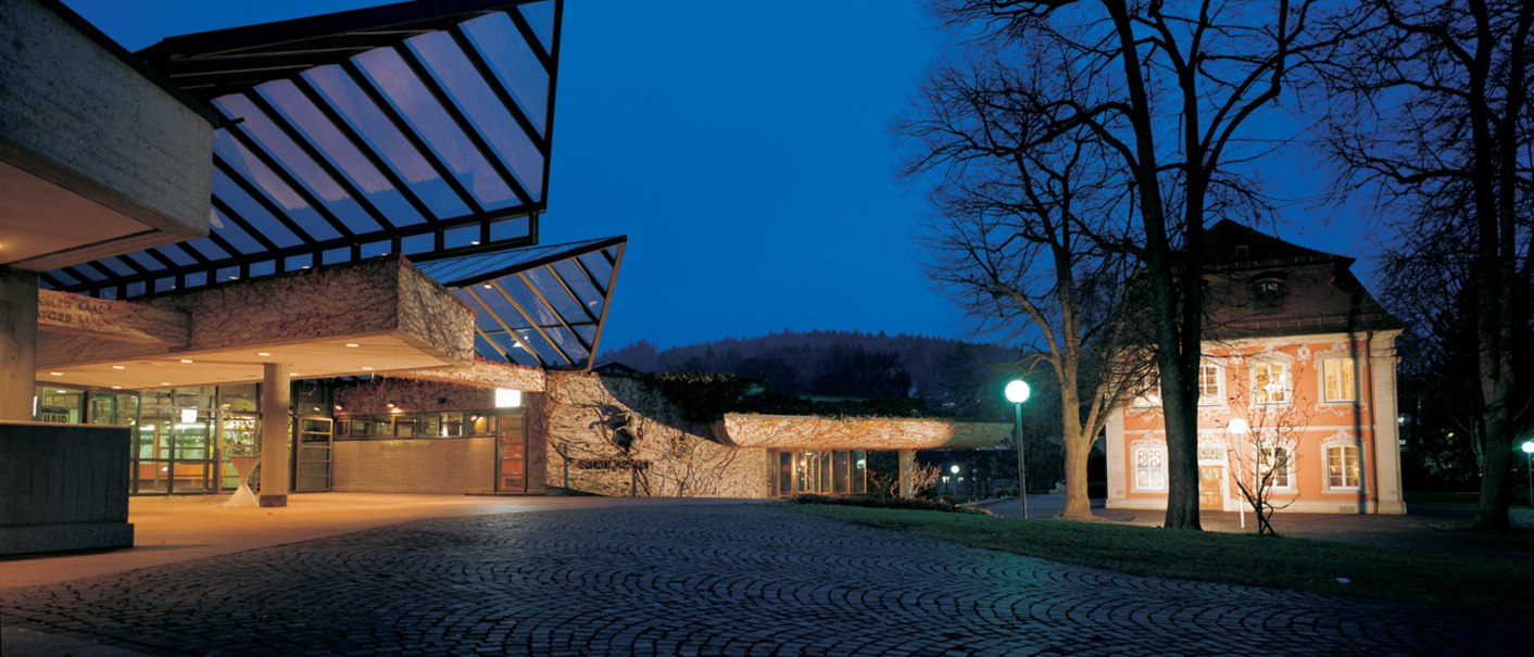 Moderne Architektur mit Glasdach bei Nacht, daneben ein beleuchtetes historisches Gebäude. Kopfsteinpflaster im Vordergrund, Bäume im Hintergrund., © Congress-Centrum Stadtgarten Schwäbisch Gmünd