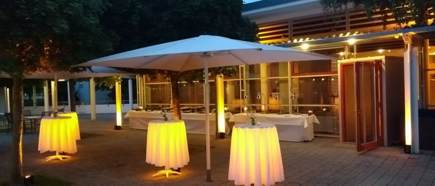Illuminated bar tables with white cloths on a terrace at dusk, surrounded by trees and a glass-fronted building., © Filharmonie_gf_1