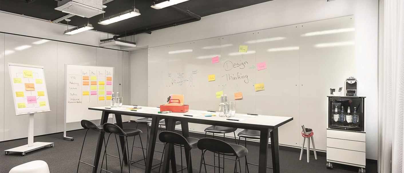 Moderner Meetingraum mit hohen Tischen, Stühlen und Whiteboards, dekoriert mit bunten Haftnotizen. Ein Flipchart und eine Kaffeemaschine sind ebenfalls vorhanden., © Copysign