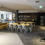 Modern lounge with a large table, white chairs and a kitchenette. Sofas and plants can be seen in the background., © INFOMOTION