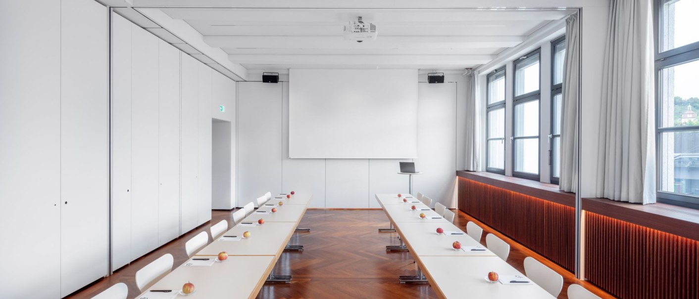 Conference room with U-shaped table arrangement, white chairs, apples and notepads on the tables, wooden floor and large windows., © © markus guhl fotografie