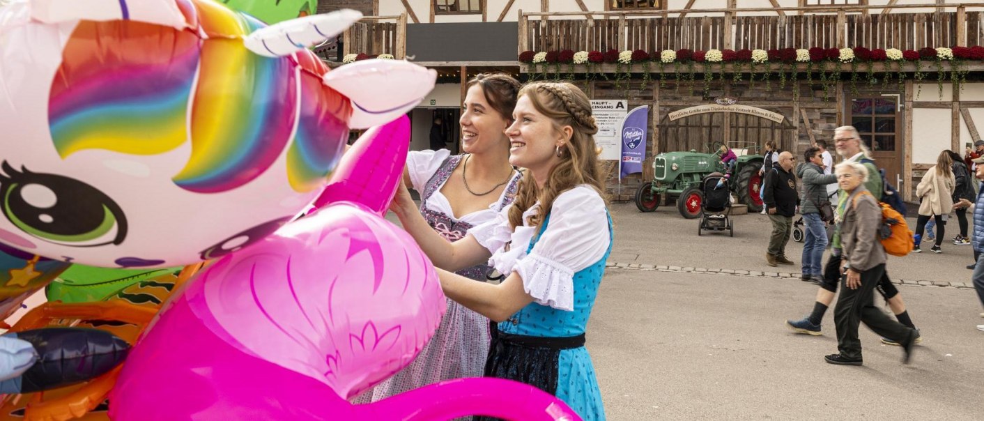 Zwei Frauen in Dirndl betrachten bunte Luftballons auf einem Volksfest in Stuttgart. Im Hintergrund sind Fachwerkhäuser und ein Traktor zu sehen., © Stuttgart-Marketing GmbH, Sarah Schmid Zwei Frauen in Dirndl betrachten bunte Luftballons auf einem Volksfest in Stuttgart. Im Hintergrund sind Fachwerkhäuser und ein Traktor zu sehen., © Stuttgart-Marketing GmbH, Sarah Schmid