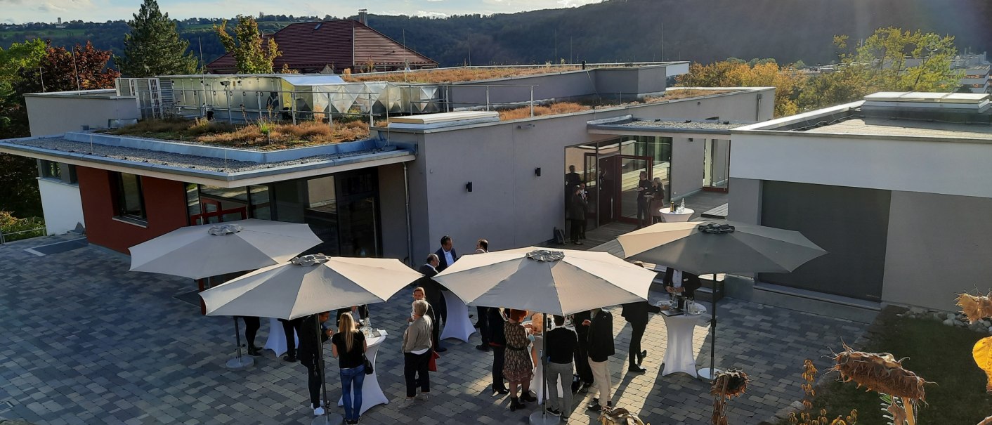 Menschen stehen unter Sonnenschirmen im Außenbereich eines modernen Gebäudes mit begrüntem Dach. Im Hintergrund sind Hügel und Bäume zu sehen., © Weingärtner Esslingen eG Menschen stehen unter Sonnenschirmen im Außenbereich eines modernen Gebäudes mit begrüntem Dach. Im Hintergrund sind Hügel und Bäume zu sehen., © Weingärtner Esslingen eG