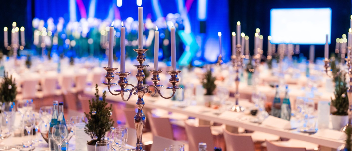 Elegante Tischdekoration mit Kerzenleuchtern, Gläsern und kleinen Tannenbäumen bei einer festlichen Veranstaltung in einer beleuchteten Arena., © Carl Benz Arena