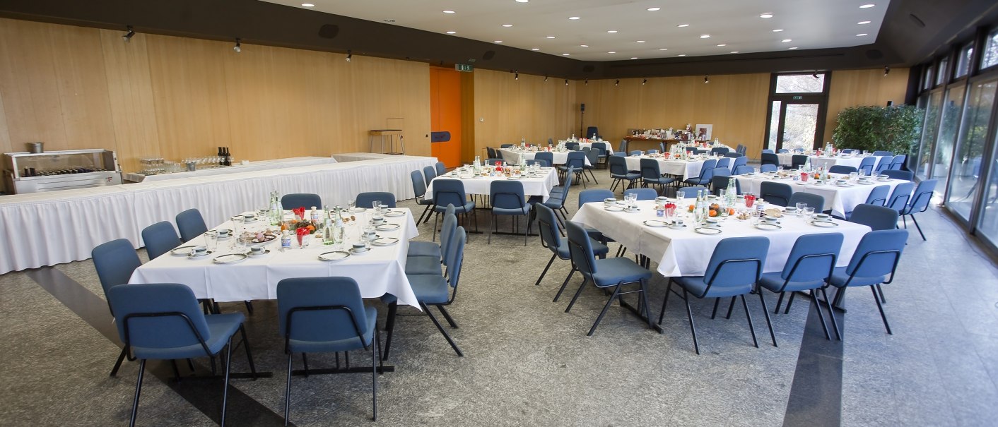 Ein festlich gedeckter Saal mit runden Tischen, weißen Tischdecken und blauen Stühlen. Im Hintergrund ein Buffetbereich., © eslive Ein festlich gedeckter Saal mit runden Tischen, weißen Tischdecken und blauen Stühlen. Im Hintergrund ein Buffetbereich., © eslive