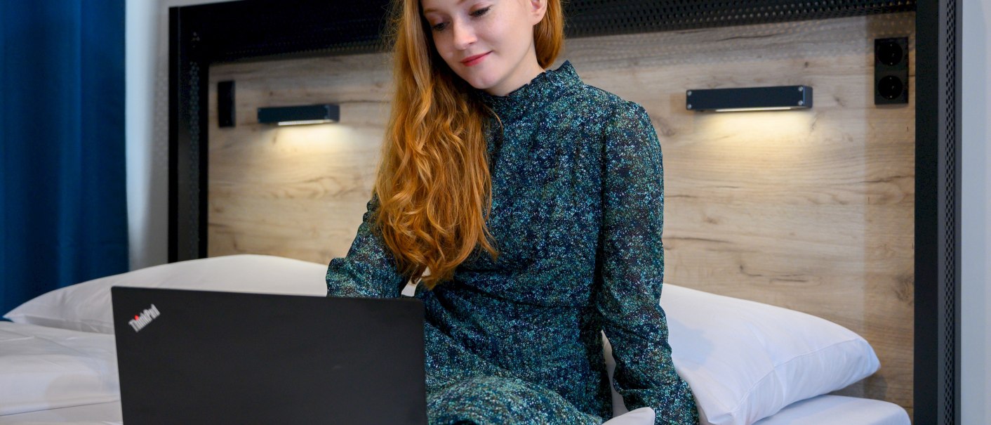 Eine Frau mit roten Haaren sitzt auf einem Bett in einem Hotelzimmer und arbeitet an einem Laptop., © a&o Hostels