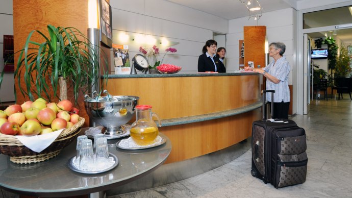A hotel reception desk with two employees helping an elderly woman with a suitcase. A table with apples and drinks is in the foreground., © Hotel Unger