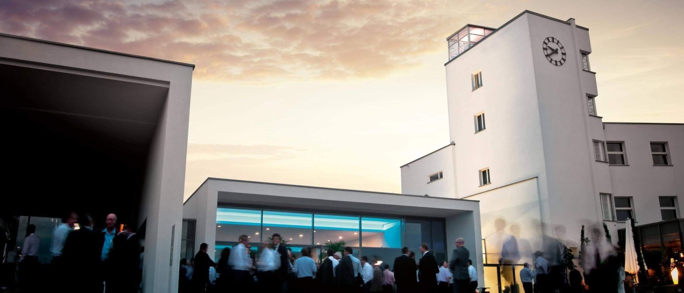 People gather in front of a modern building with a clock tower while the sky glows in the sunset., © rauschenbergercatering