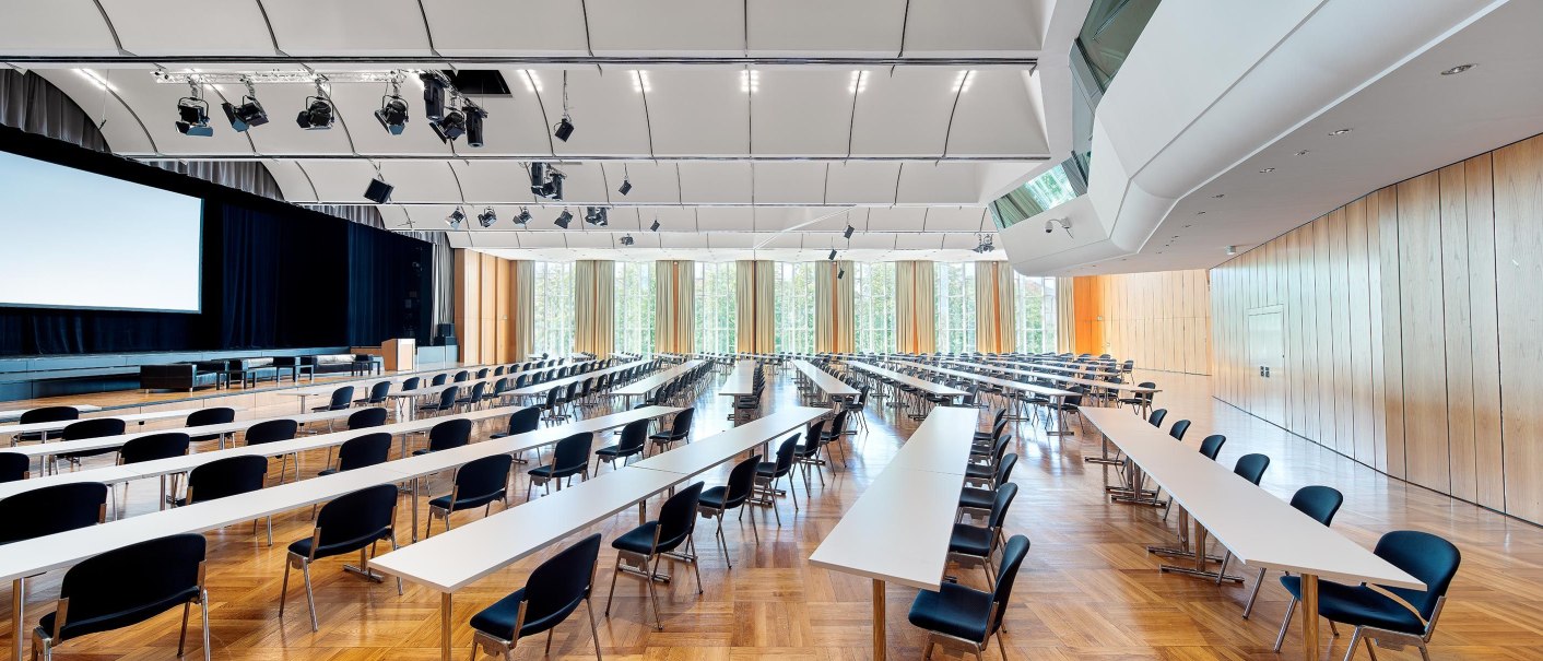 Modern conference room with long tables and chairs, large stage and screen. Bright window front and wood paneling., © Stauch