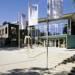 Modern hall with glass façade and flags in the foreground, surrounded by trees and blue sky., © Barbara-Künkelin-Halle