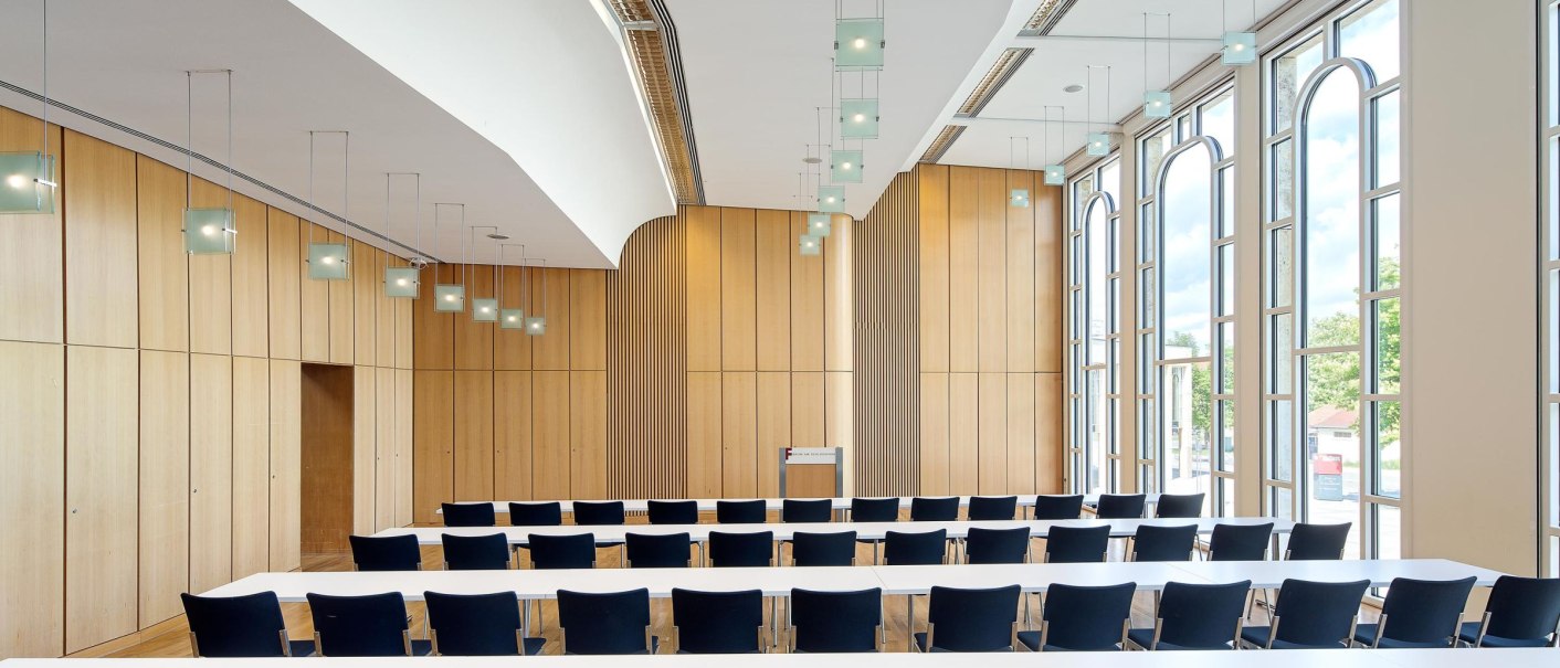 Modern conference room with wood paneling, large windows and hanging lights. Rows of black chairs and white tables are arranged., © Stauch
