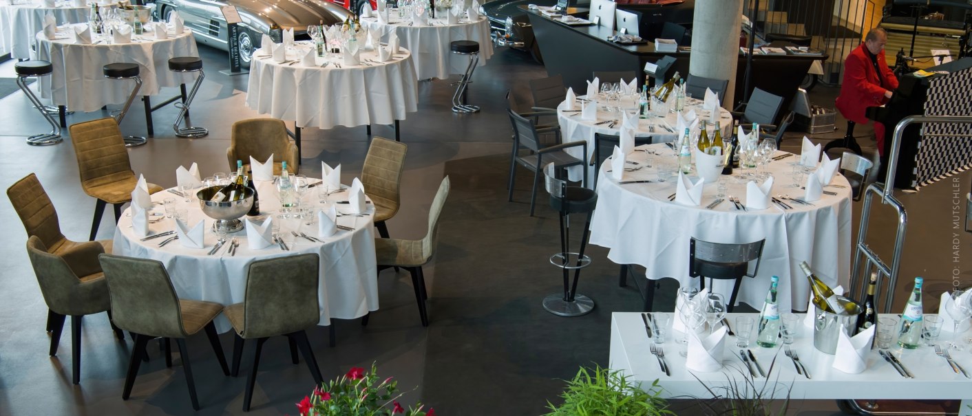 Eleganter Bankettsaal im V8 HOTEL mit gedeckten Tischen und Oldtimern im Hintergrund. Ein Pianist spielt im roten Anzug., © Hardy Mutschler
